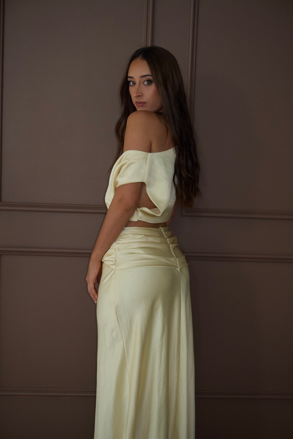 A woman wearing a cream satin two-piece outfit consisting of an off-the-shoulder crop top and a matching high-waisted pleated maxi skirt, posing against a brown paneled wall.