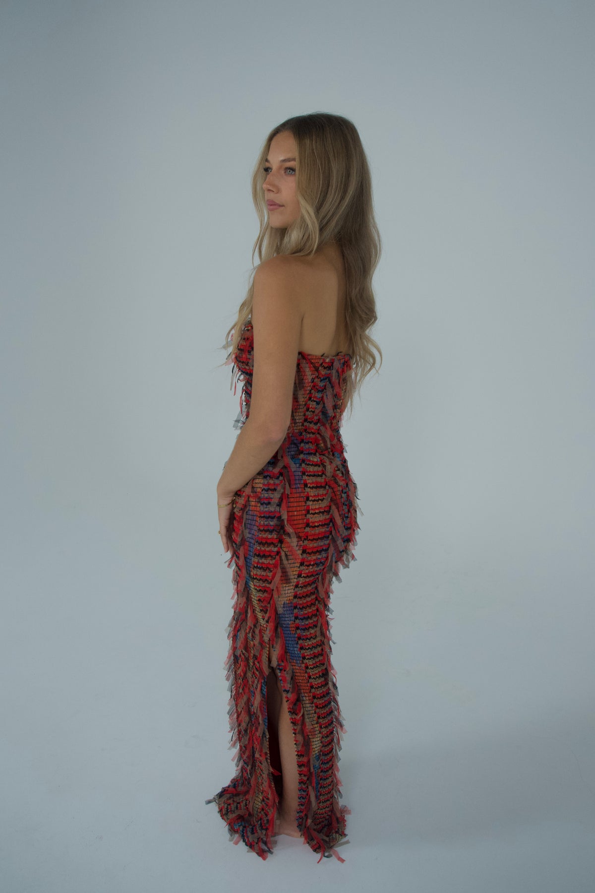 Side profile of a woman wearing a strapless, floor-length textured maxi dress featuring layered fringe detailing and a multicoloured red, black, purple and neutral woven pattern, photographed against a light studio backdrop.