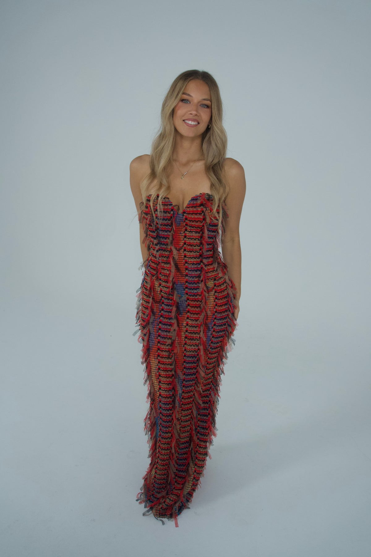 A woman modelling a strapless floor-length gown featuring a vibrant red, black and multicoloured woven pattern with textured fringe detailing throughout and a fitted silhouette, photographed against a light grey studio backdrop.