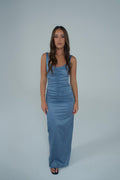 A woman modeling a sleeveless, floor-length blue satin gown with ruched detailing on the bodice, standing against a solid light gray studio background. - @sarahmadden