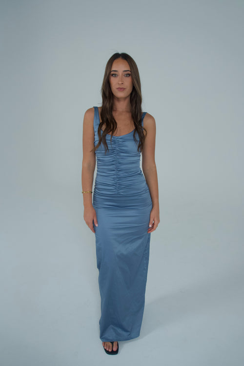 A woman modeling a sleeveless, floor-length blue satin gown with ruched detailing on the bodice, standing against a solid light gray studio background. - @sarahmadden