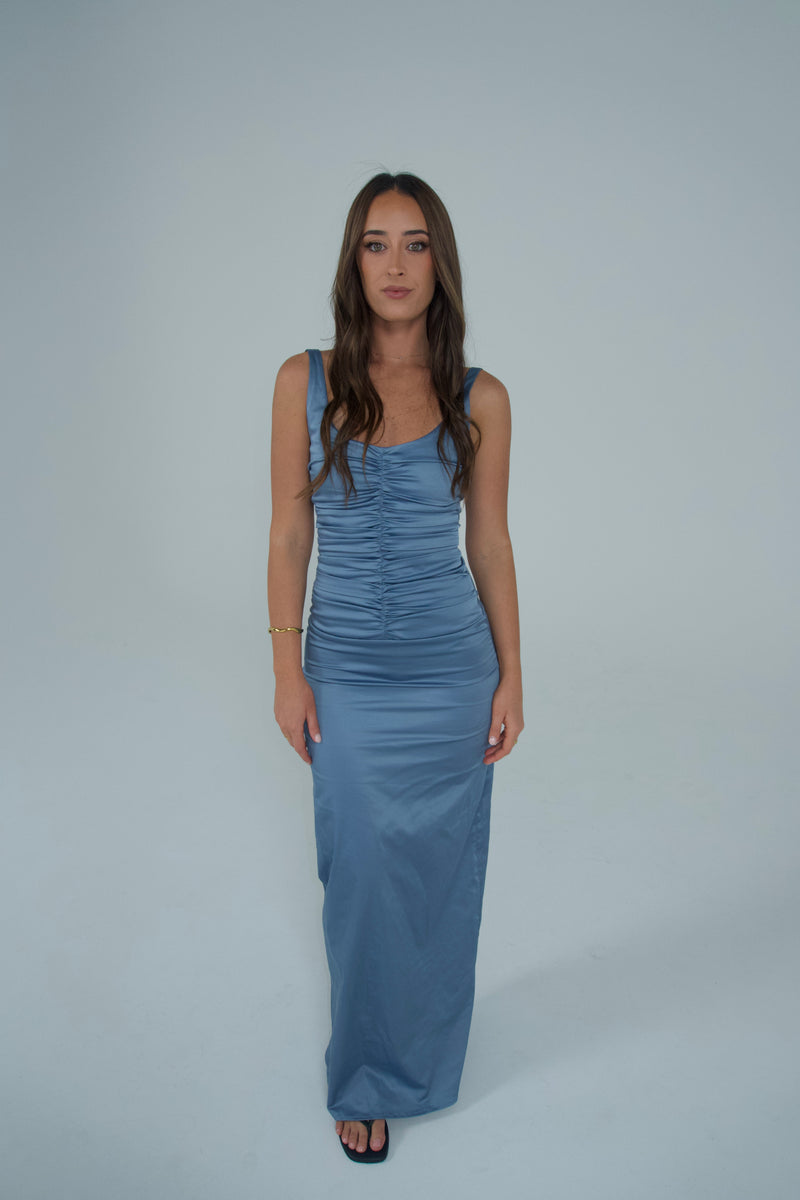 A woman modeling a sleeveless, floor-length blue satin gown with ruched detailing on the bodice, standing against a solid light gray studio background. - @sarahmadden