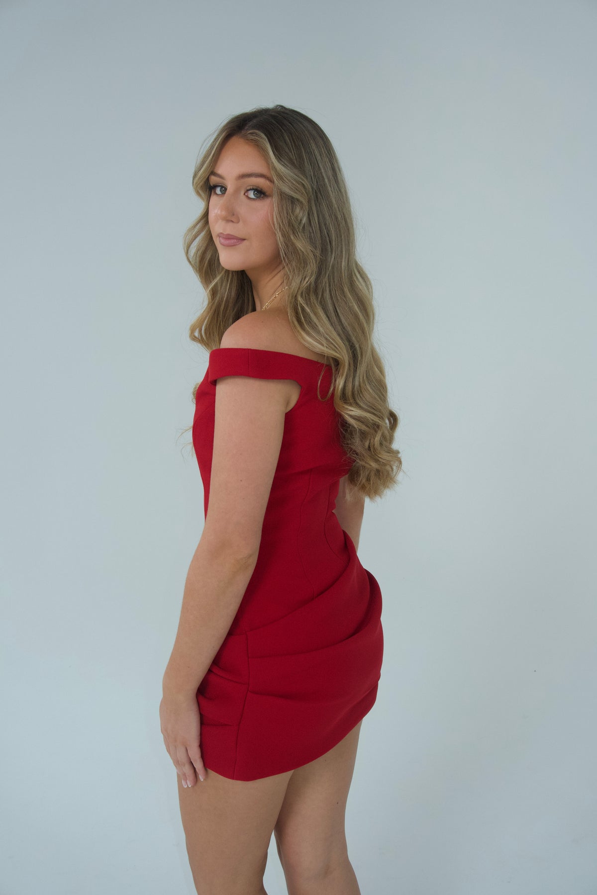A woman with long wavy blonde hair models an off-the-shoulder red mini dress against a plain light gray studio background.