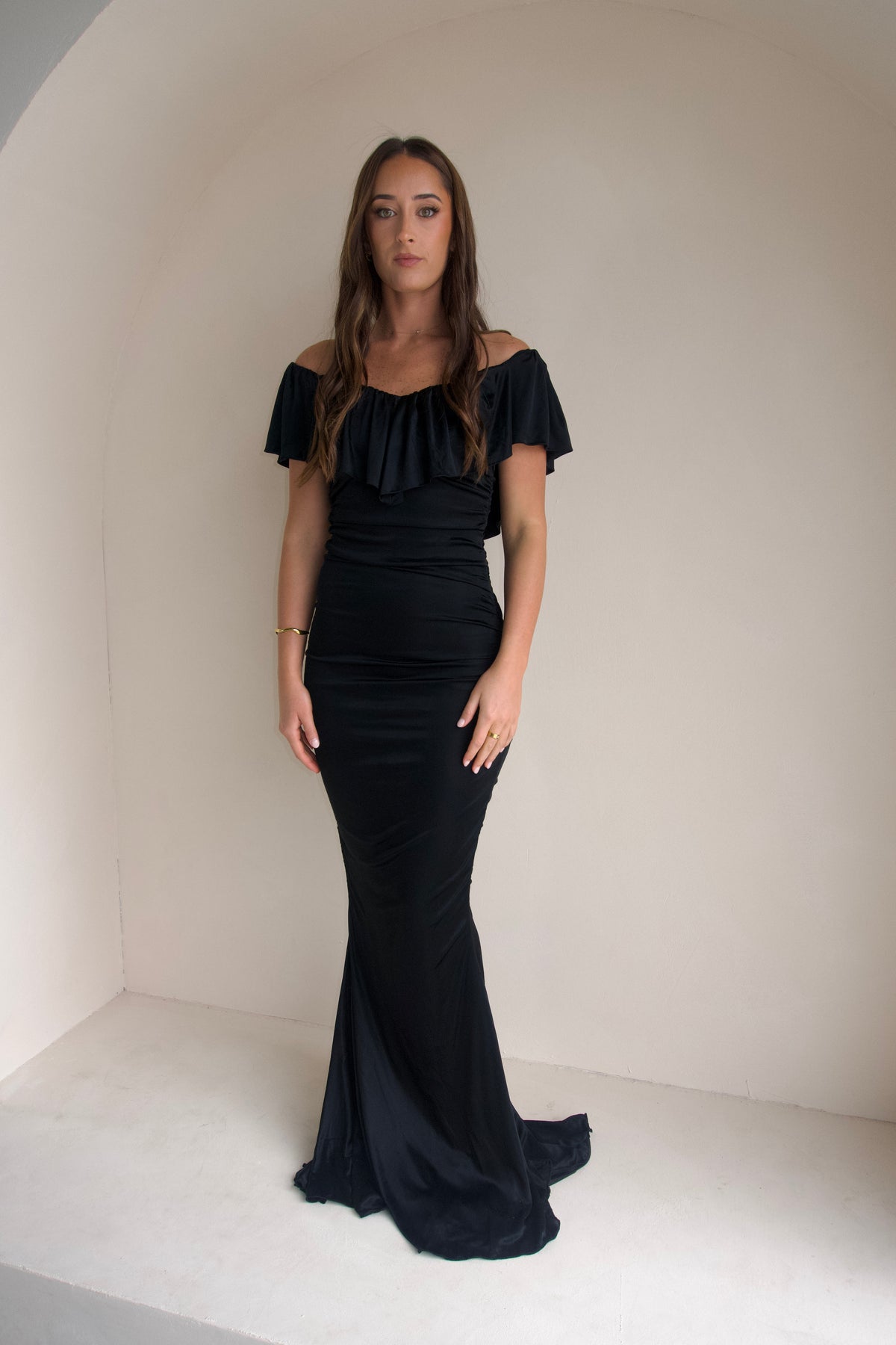 Woman wearing a black floor-length fitted gown with an off-the-shoulder ruffled neckline and softly ruched silhouette, photographed against a neutral cream studio background.