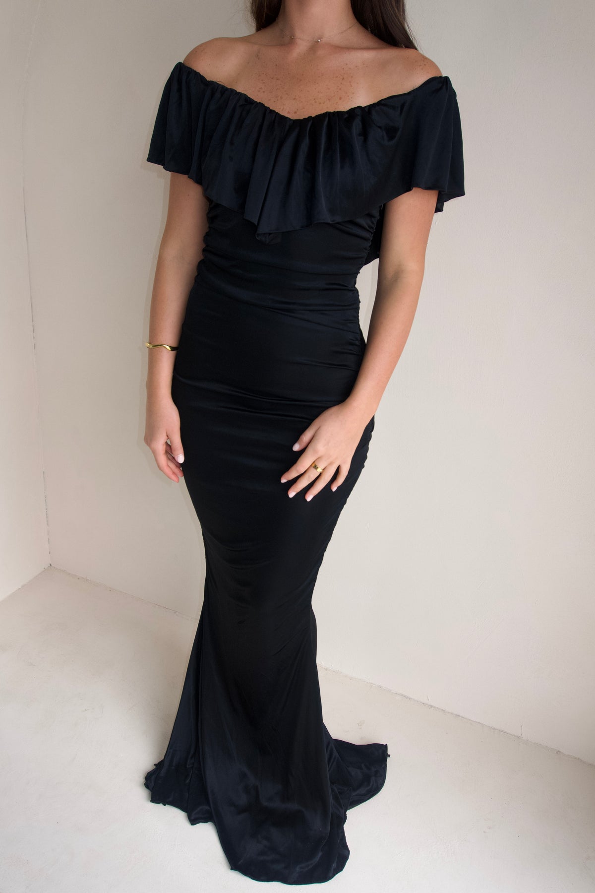 Close up Women wearing a black floor-length fitted gown with an off-the-shoulder ruffled neckline and softly ruched silhouette, photographed against a neutral cream studio background.