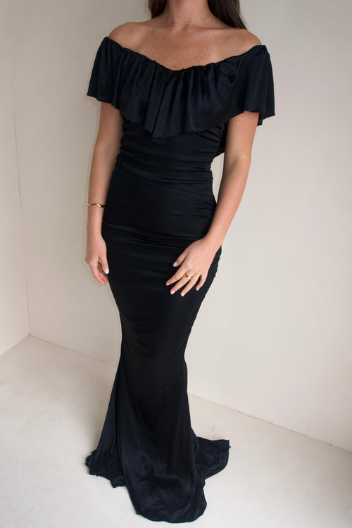 Close up Women wearing a black floor-length fitted gown with an off-the-shoulder ruffled neckline and softly ruched silhouette, photographed against a neutral cream studio background.