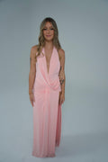 Woman modelling a soft pink floor-length halter gown featuring a deep plunging neckline, draped waist detailing and a thigh-high side split, photographed front-on against a soft grey studio background.