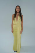 Front view of a woman wearing a pale yellow halter-neck maxi dress with a plunging neckline, ruched waist and long draped sash detail, photographed against a light studio backdrop.