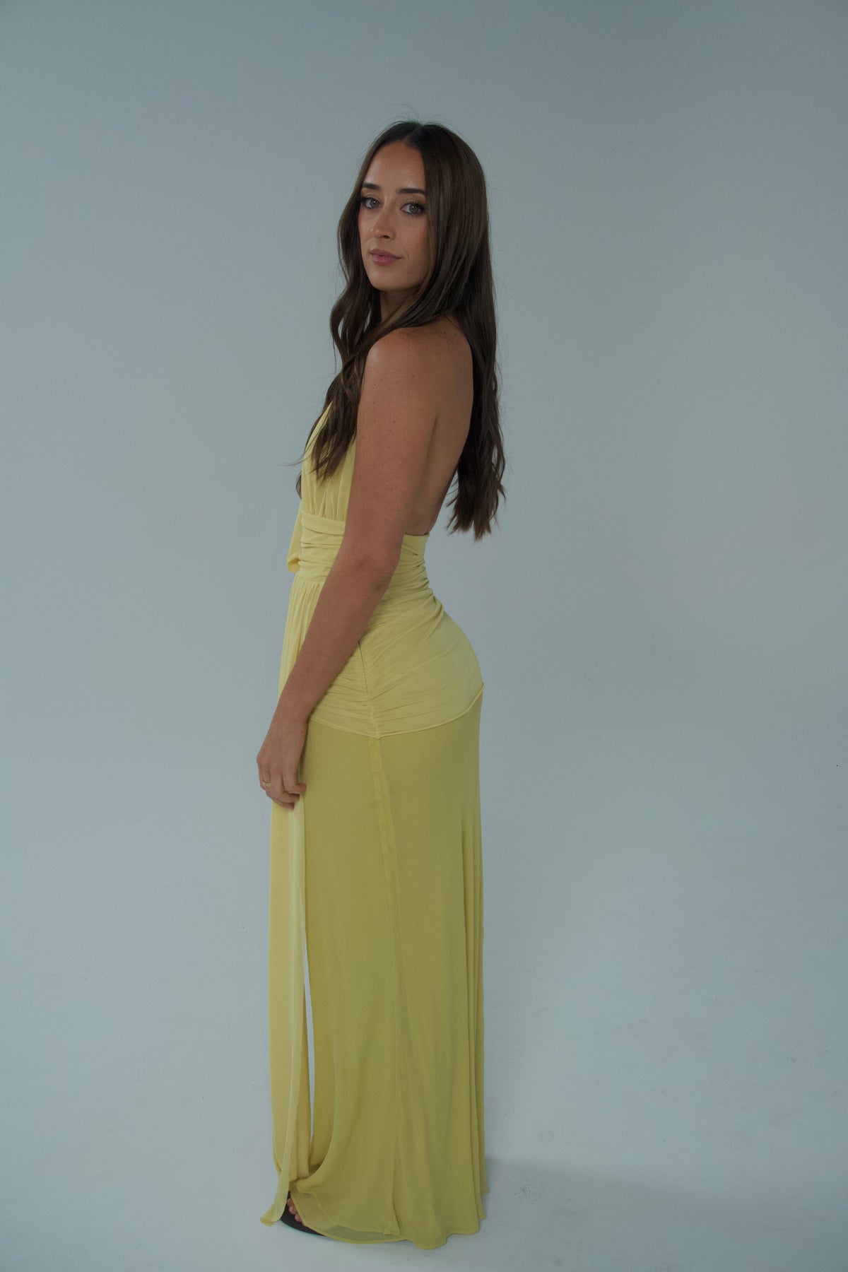 Side view of a woman wearing a pale yellow halter-neck maxi dress with an open back, ruched waist and long flowing draped sash, photographed against a light studio backdrop.