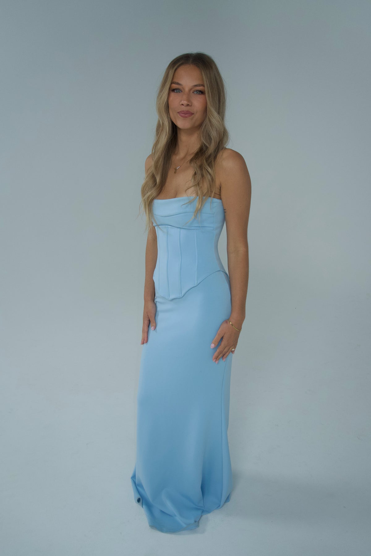 Front view of a woman wearing a strapless powder blue floor-length gown with a structured corset-style bodice, fitted silhouette and softly flared hem, photographed against a light studio backdrop.