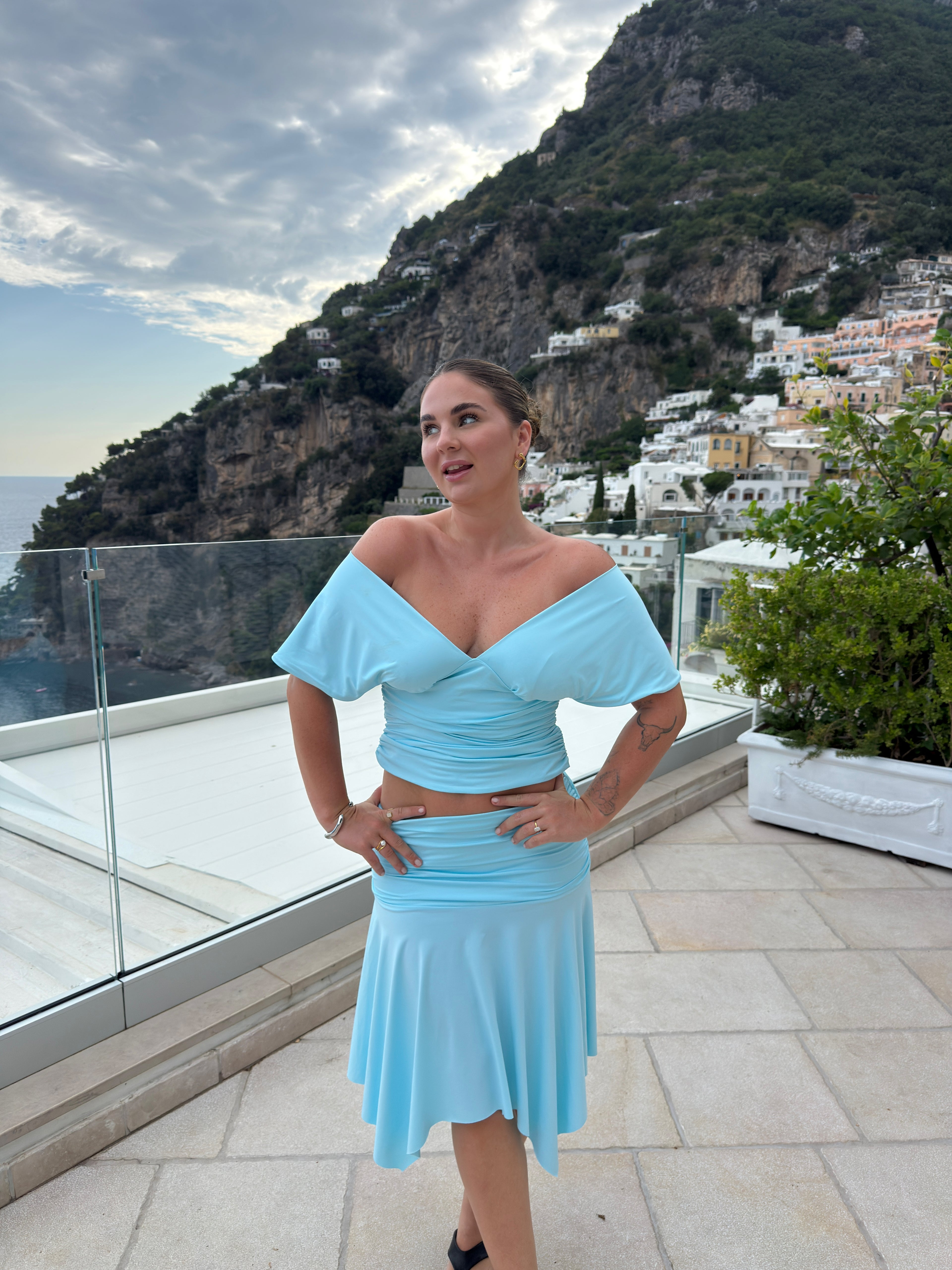 A woman modeling a light blue two-piece dress set consisting of an off-the-shoulder crop top and a matching flared skirt, standing on a terrace with a scenic coastal mountain and white buildings in the background.