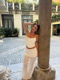 A woman posing outdoors in a white two-piece crochet knit set consisting of a crop top and a long skirt, leaning against a stone column in a courtyard.