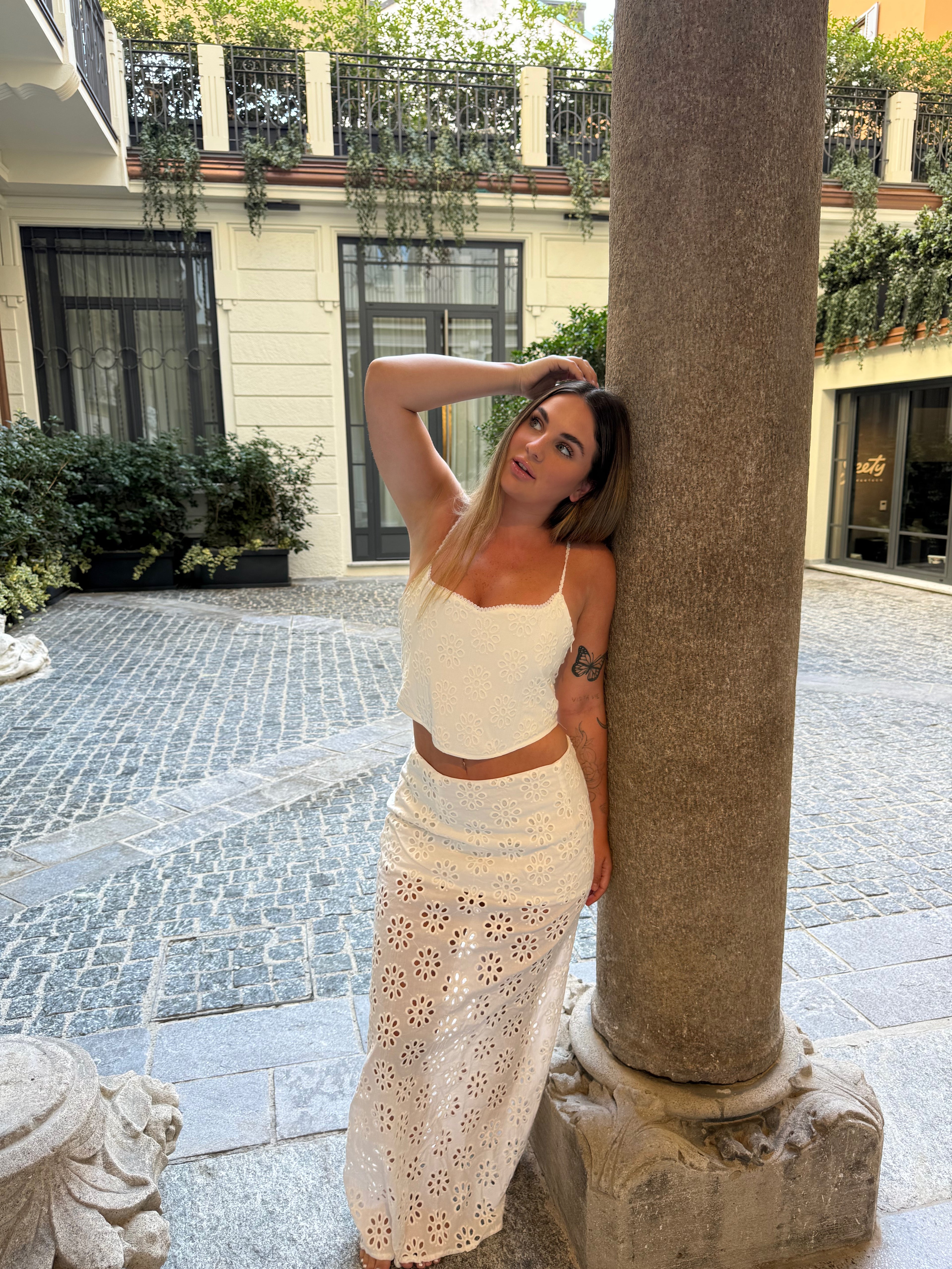 A woman posing outdoors in a white two-piece crochet knit set consisting of a crop top and a long skirt, leaning against a stone column in a courtyard.