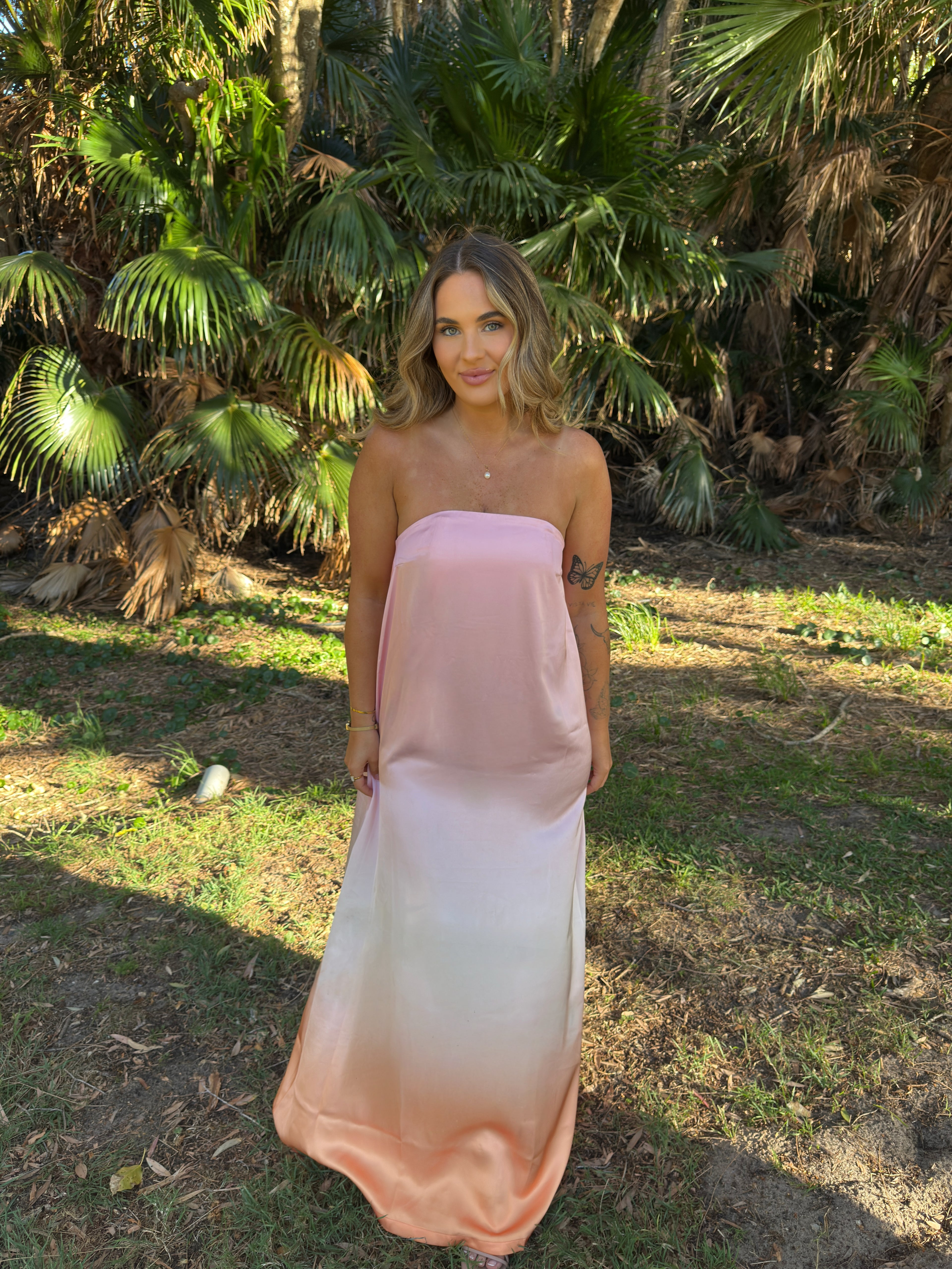 Woman wearing a pink strapless dress standing in a natural setting with palm trees. - @chloecid