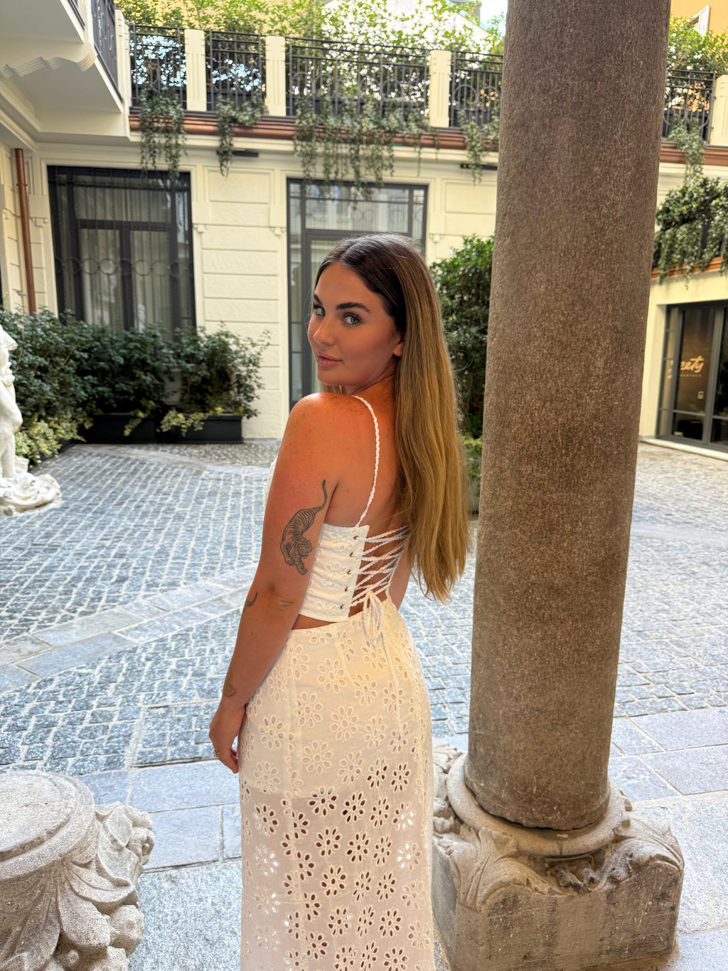 A woman wearing a white eyelet lace dress with a corset-style open back, standing in an outdoor courtyard next to a large stone column.