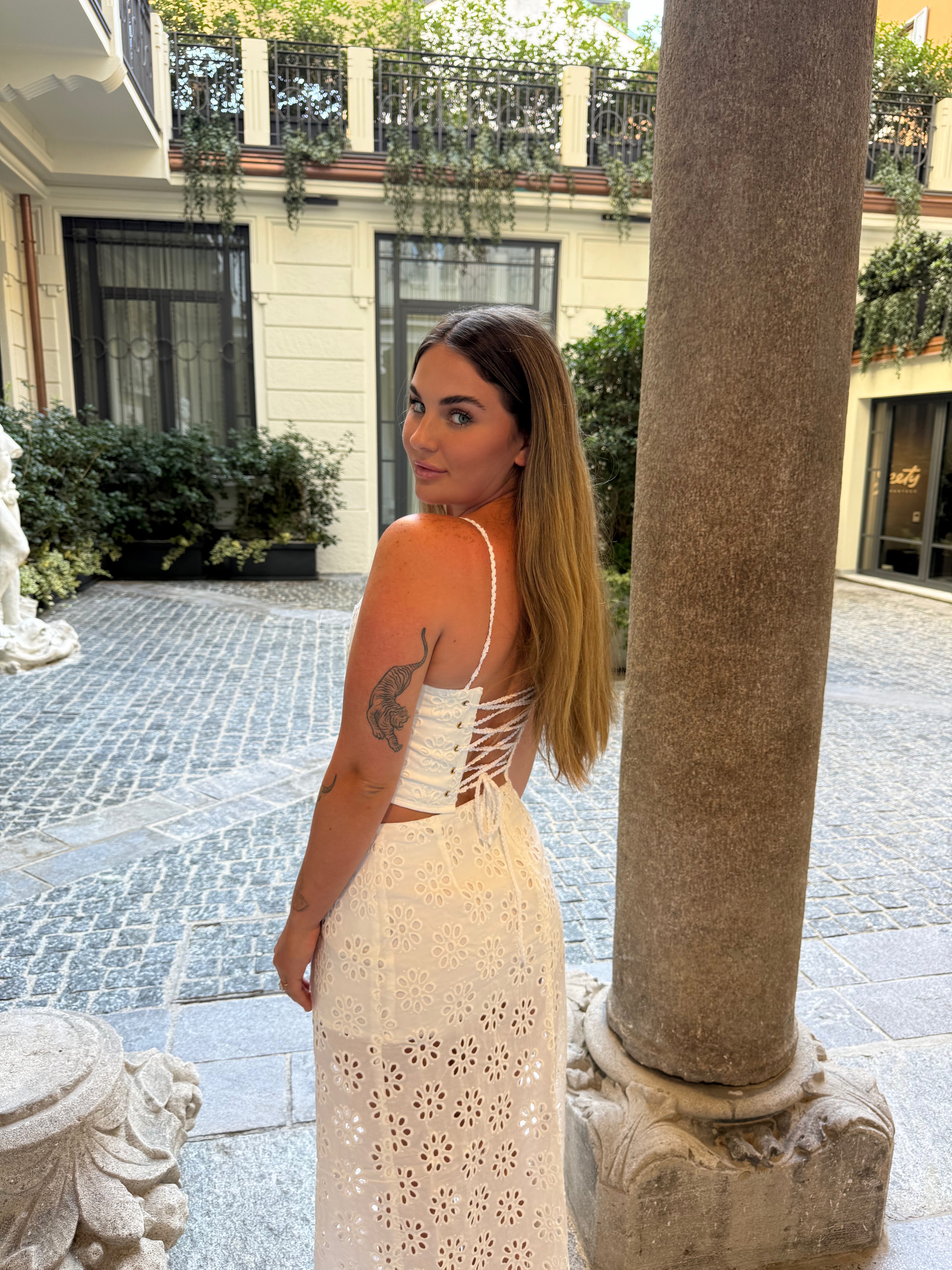 A woman wearing a white eyelet lace dress with a corset-style open back, standing in an outdoor courtyard next to a large stone column.