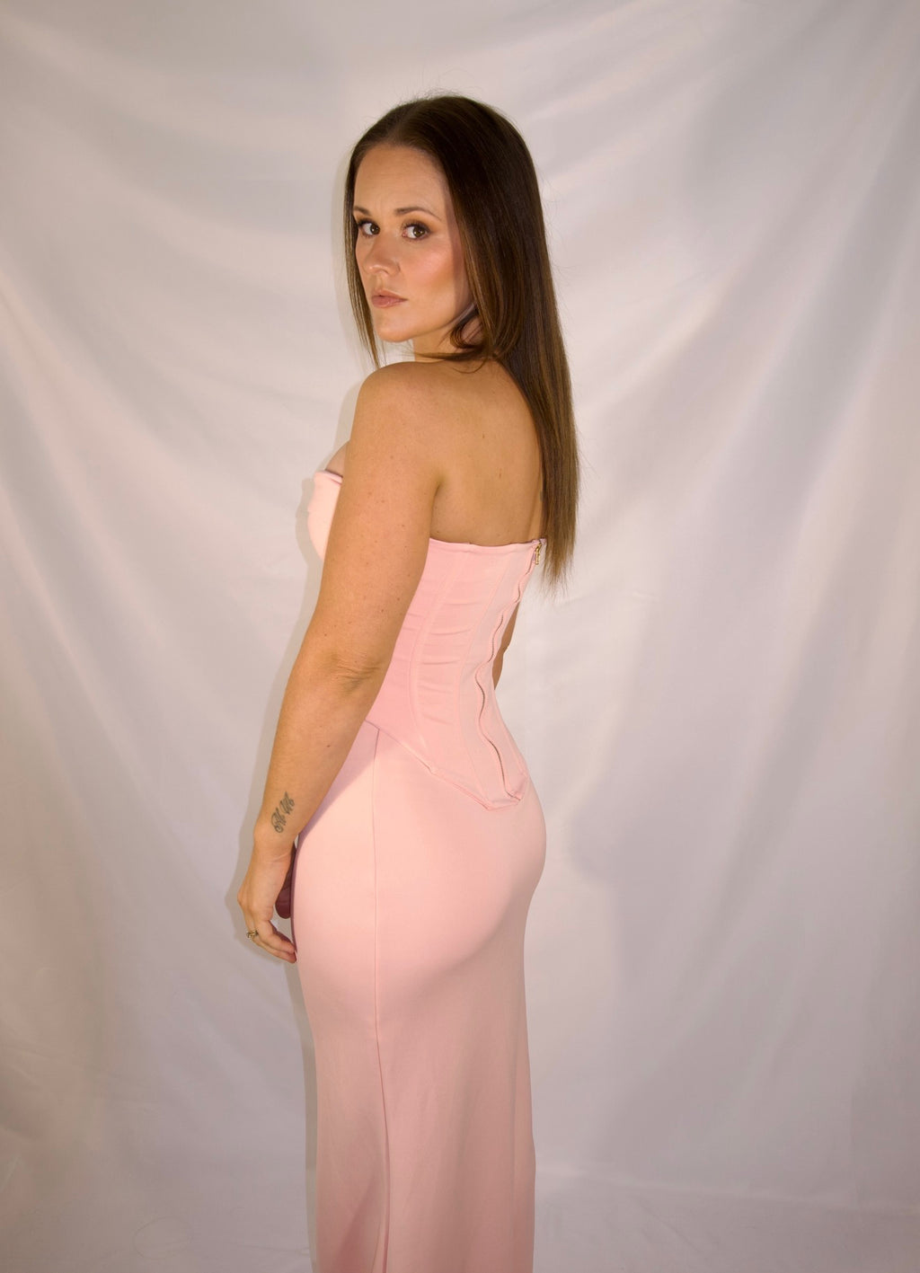 A woman modeling a strapless light pink evening gown, shown from the back against a white draped fabric backdrop.