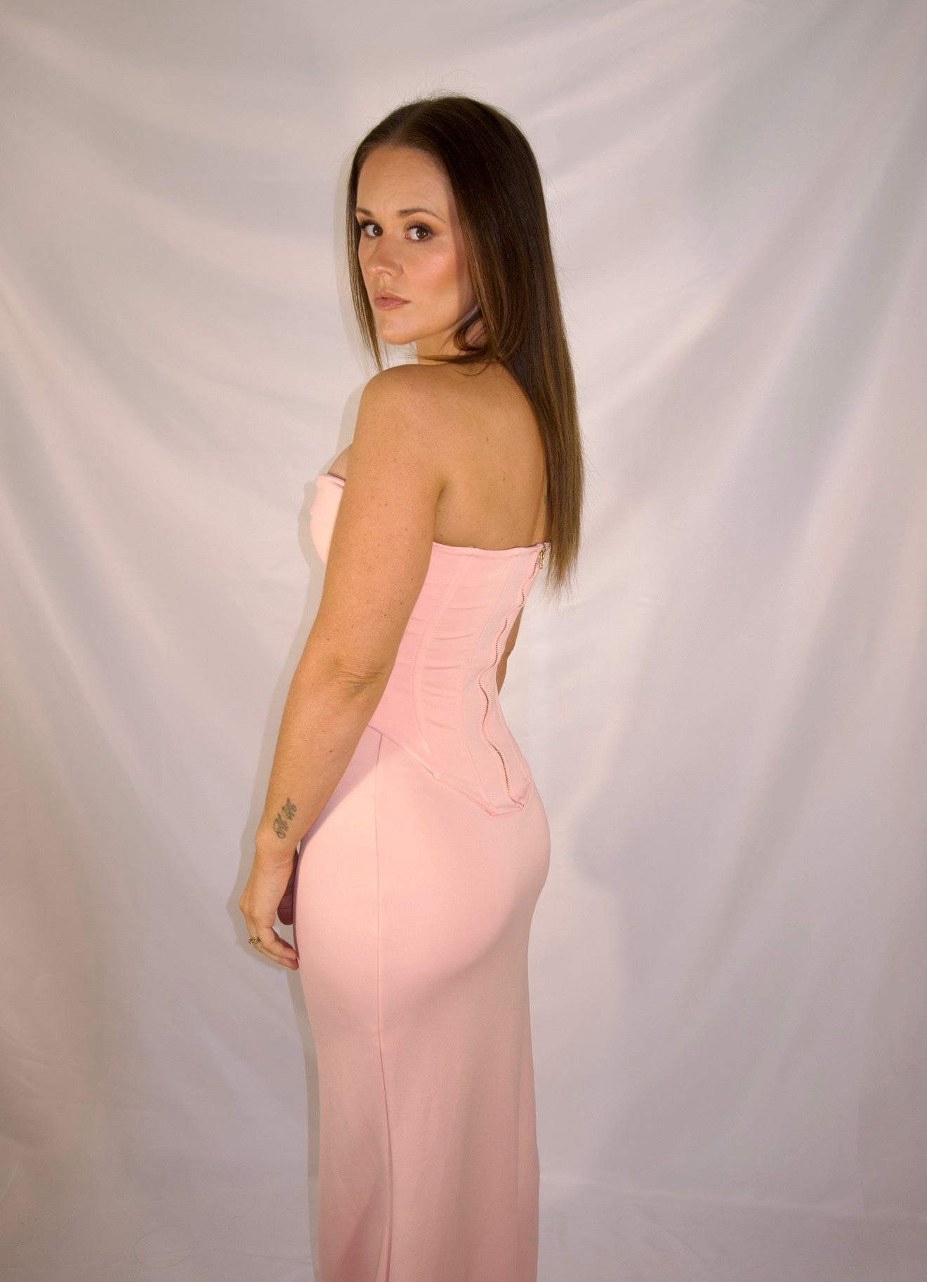 A woman modeling a strapless light pink evening gown, shown from the back against a white draped fabric backdrop.