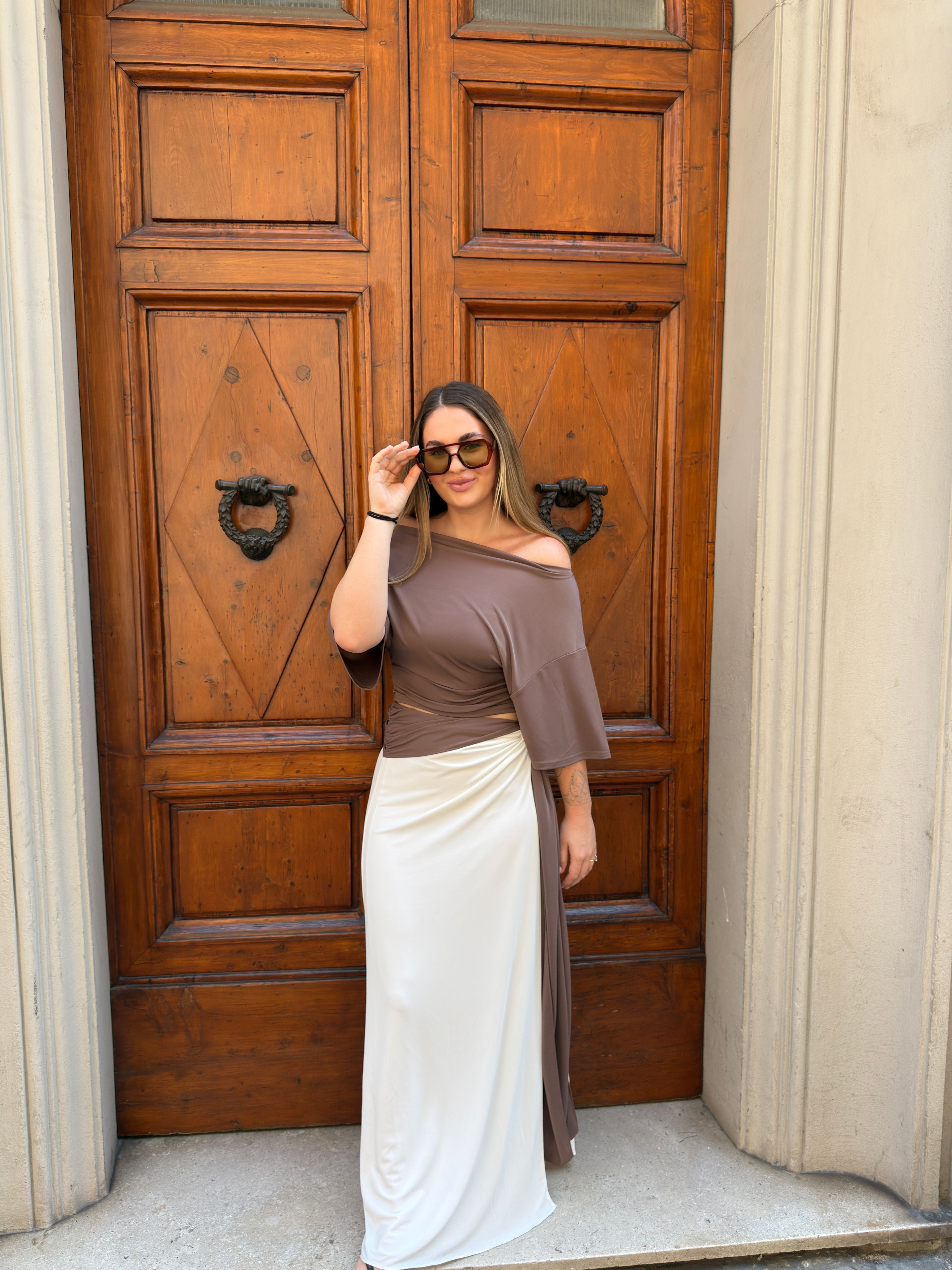 A woman wearing a taupe off-the-shoulder top and a cream wrap skirt stands in front of a large wooden door.