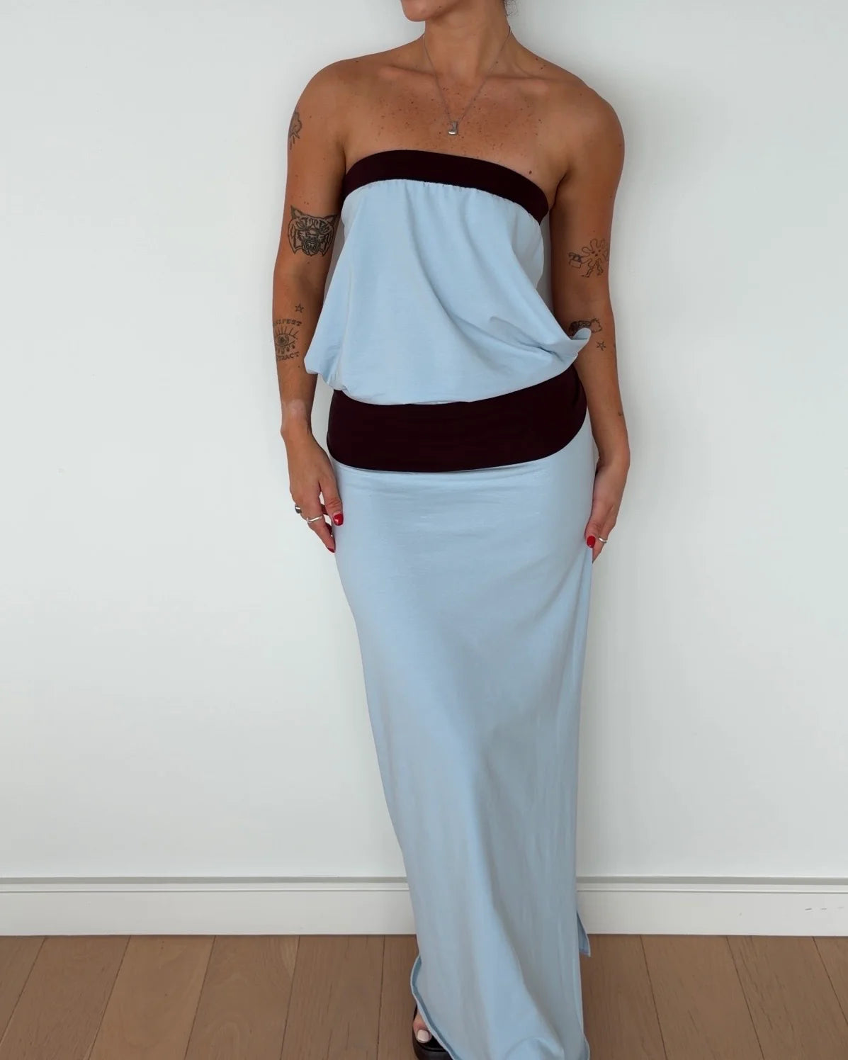 A woman modeling a strapless light blue maxi dress with contrasting dark burgundy velvet trim at the bust and waist, standing against a white wall.