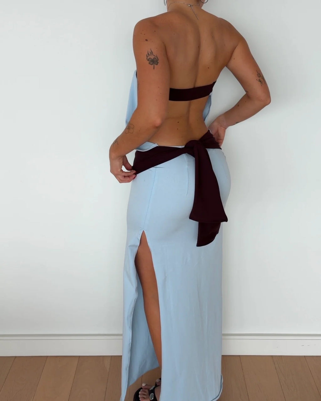 A woman shown from the back wearing a light blue maxi dress with a dark brown halter-style strap across the upper back and a matching dark brown fabric tie at the waist, standing against a plain white wall.