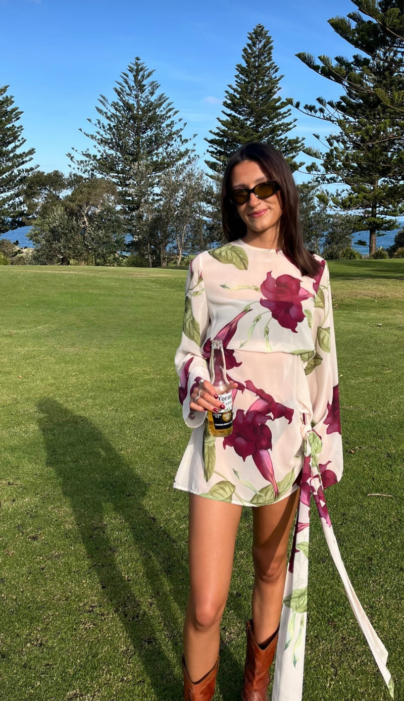 Woman taking a mirror selfie wearing the By Nicola Elodie Mini Dress, featuring a cream base with burgundy floral print, green foliage, long sleeves and a draped side tie with a flowing floor-length panel. @ellahbates