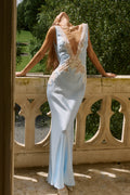 A woman wearing a light blue satin evening gown with lace and beaded embellishments, posing on a stone balcony with a garden background.