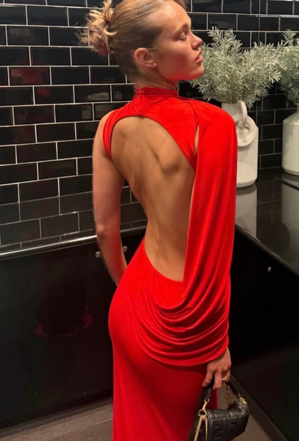 A woman wearing a bright red backless evening gown with draped fabric details, standing in front of a black tiled wall.