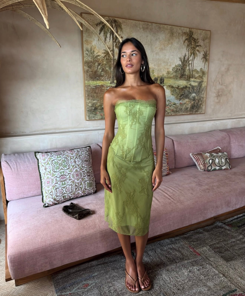 A woman modeling a strapless olive green midi dress with a structured corset-style bodice and sheer overlay, standing in a living room in front of a pink sofa.
