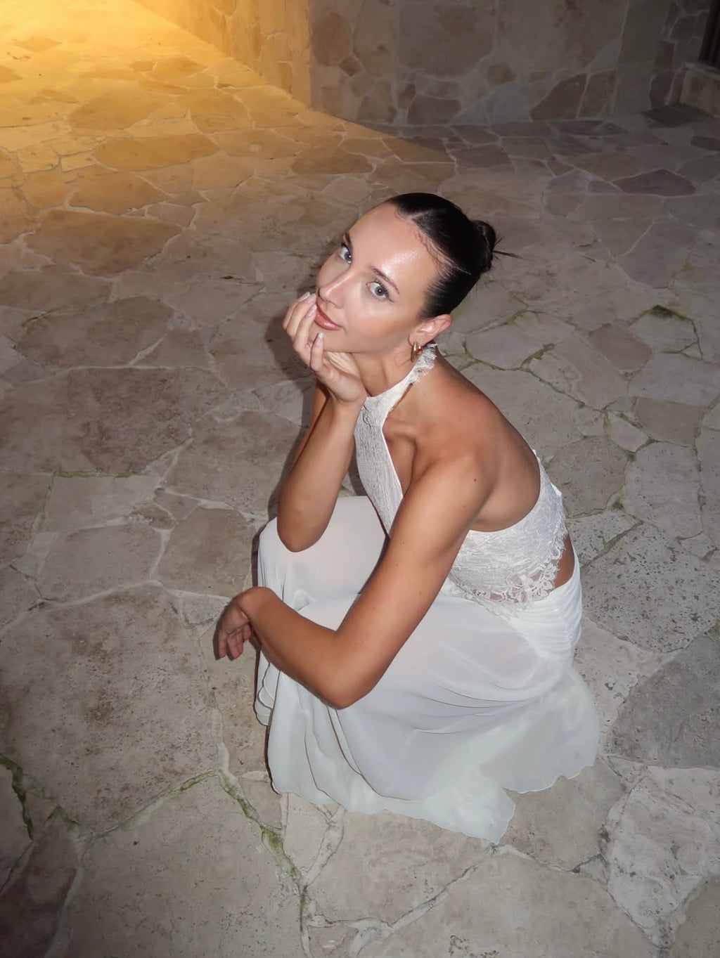 A woman in a white halter-neck dress with lace detailing sits on a stone floor in a dimly lit stone-walled environment.