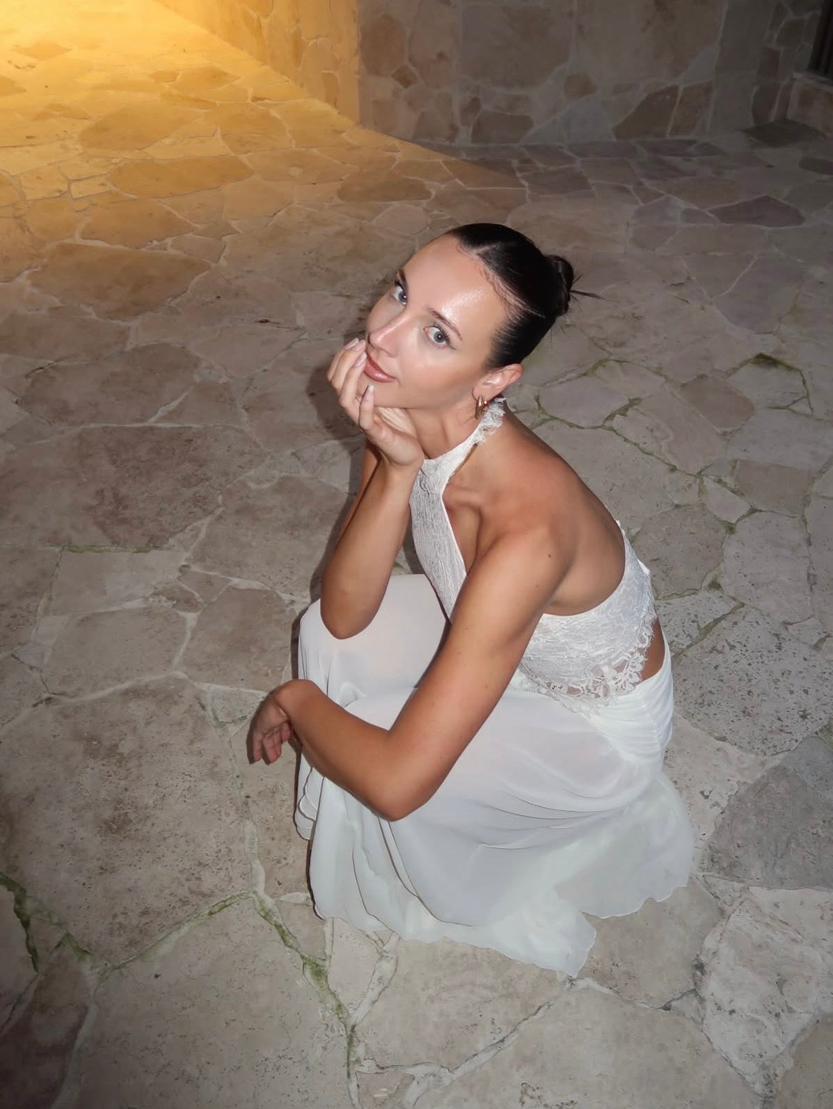 A woman in a white halter-neck dress with lace detailing sits on a stone floor in a dimly lit stone-walled environment.