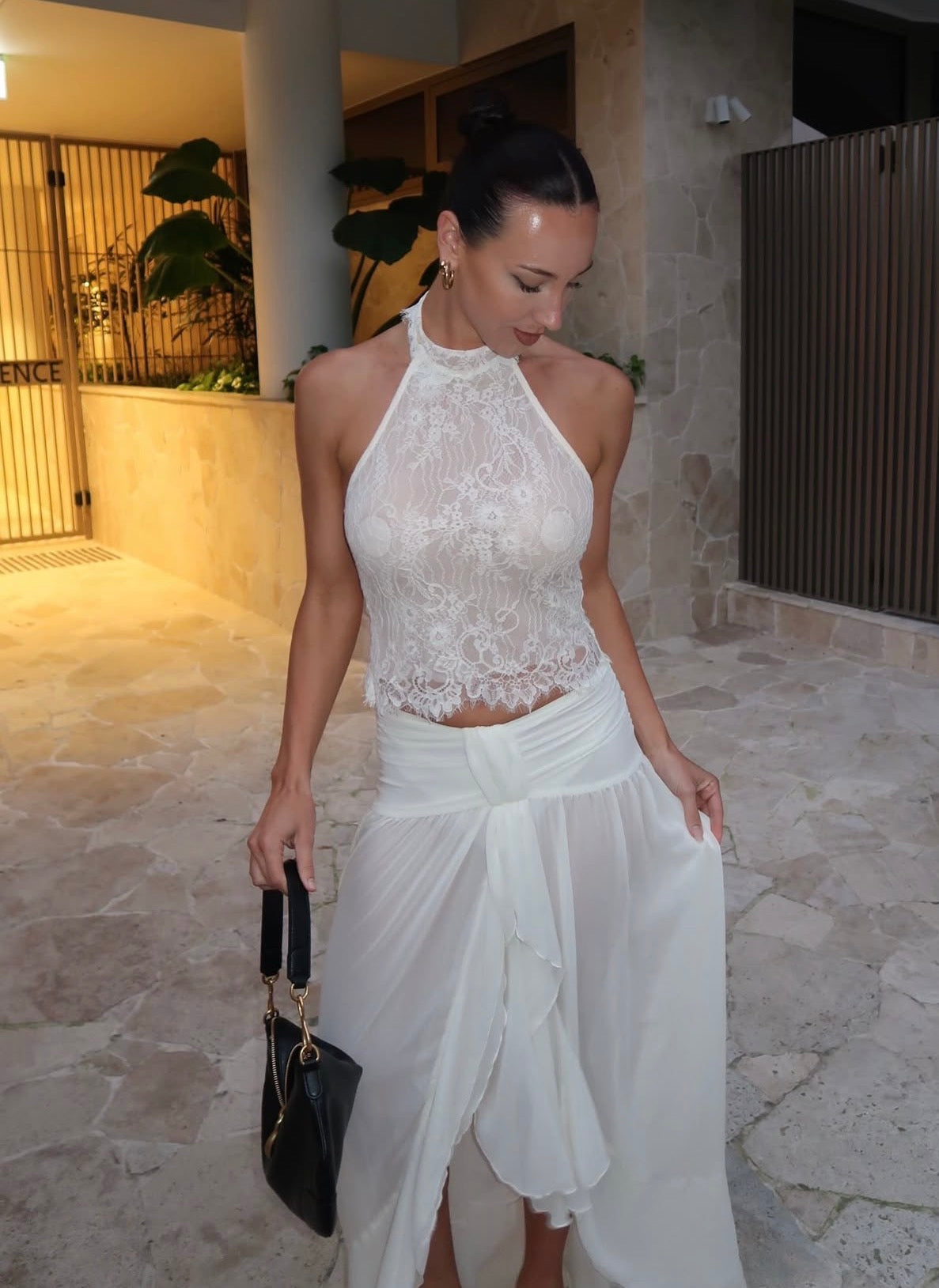 A woman wearing a white lace halter crop top and a flowing white wrap skirt, holding a black handbag in an outdoor stone walkway setting.