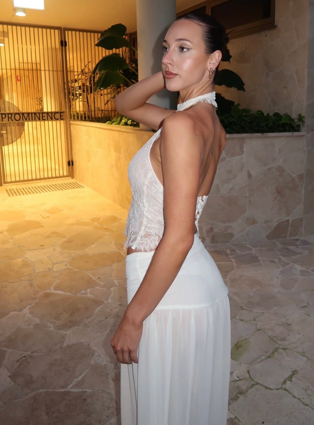A woman wearing a white sleeveless halter-neck dress with a lace bodice and flowing skirt, standing in a warm-toned stone walkway.
