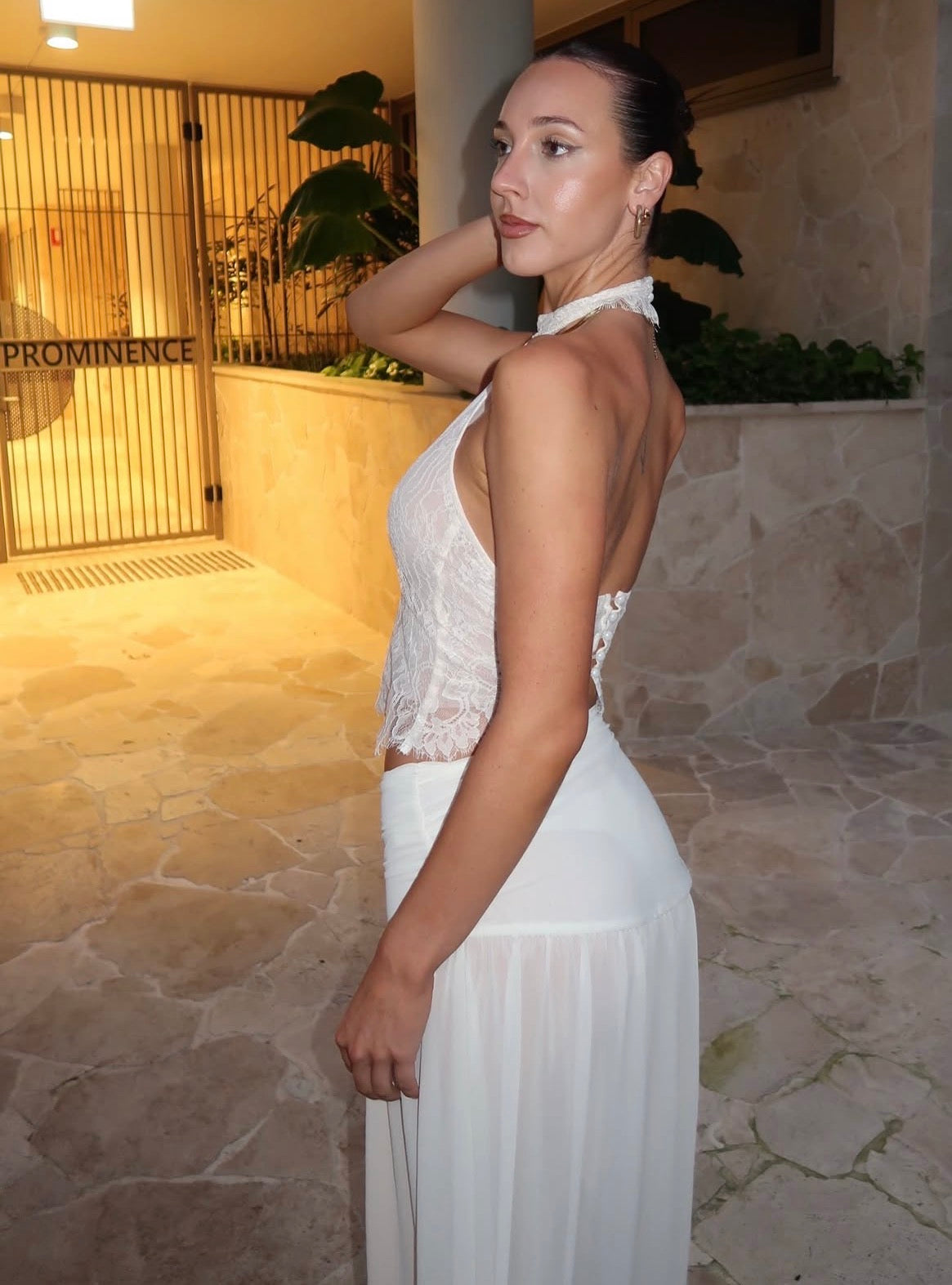 A woman wearing a white sleeveless halter-neck dress with a lace bodice and flowing skirt, standing in a warm-toned stone walkway.