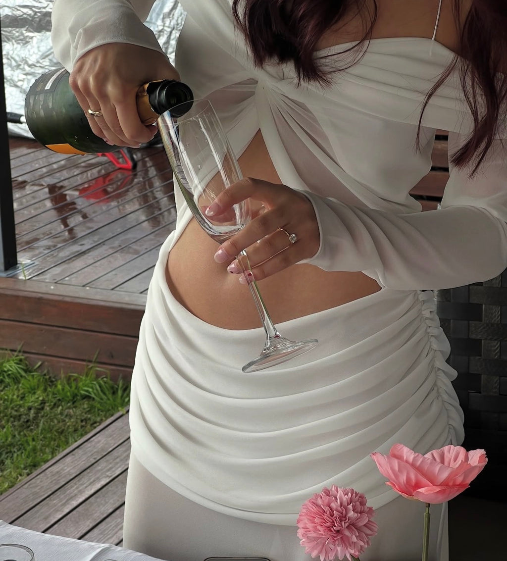 A woman wearing a white long-sleeve dress with side cutouts and a front twist detail, pouring champagne from a bottle into a glass on a wooden deck.