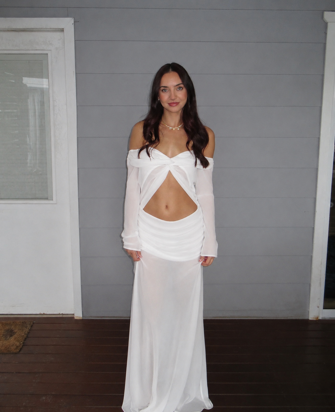 A woman modeling a white two-piece outfit consisting of an off-the-shoulder long-sleeve crop top with a twisted front detail and a matching sheer maxi skirt, standing against a gray paneled wall. - @chelseabalzan_