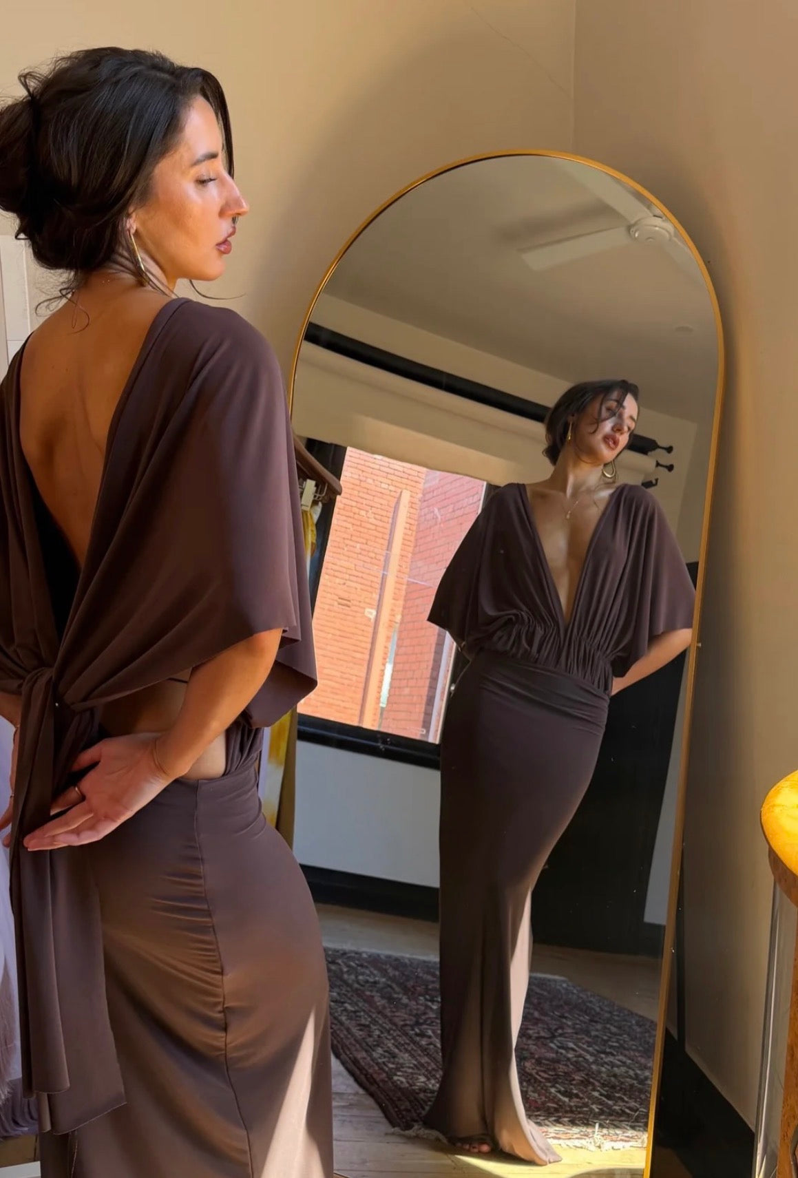 A woman wearing a dark brown draped maxi dress with a deep back and flared sleeves, standing in front of a large arched mirror that reflects her profile. The dress has a matching fitted skirt.