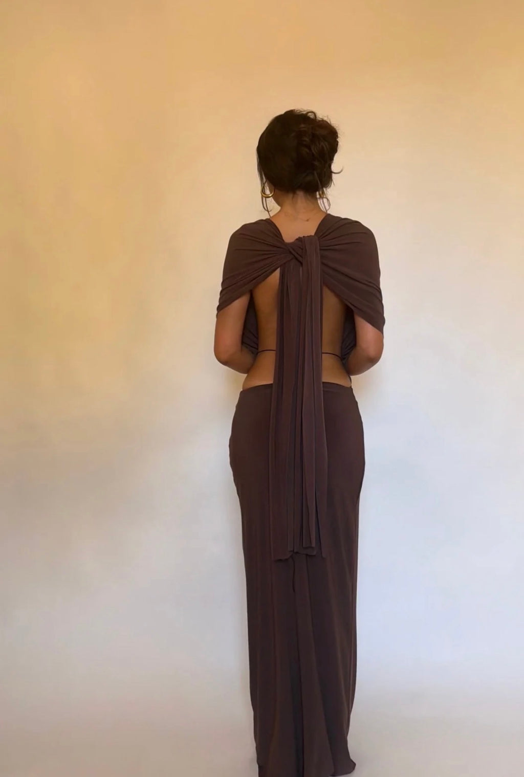 A woman stands with her back to the camera wearing a floor-length dark brown wrap dress with short draped sleeves and a large back tie, set against a plain beige wall.