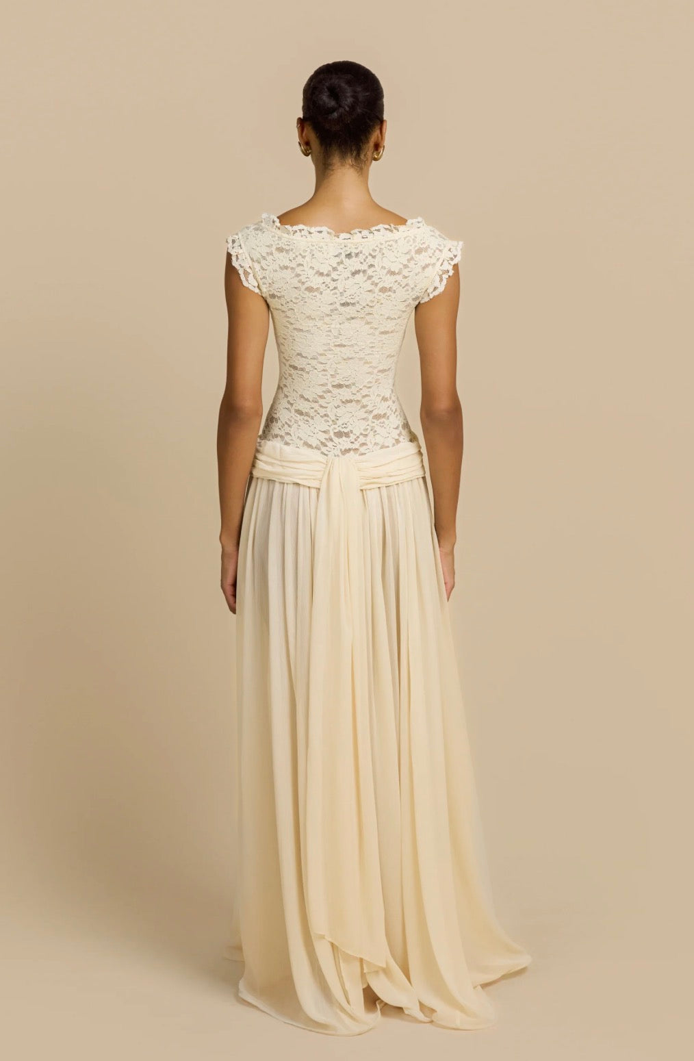 A woman shown from the back wearing a floor-length cream dress with a lace bodice, cap sleeves, and a flowing chiffon skirt against a plain beige studio background.