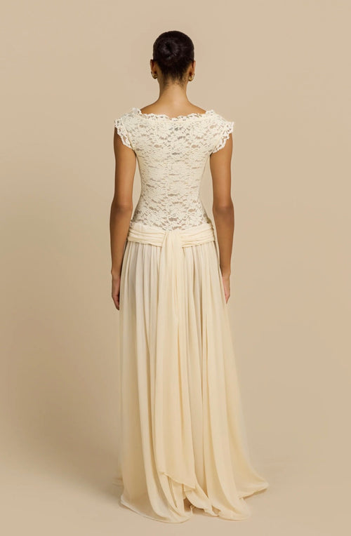 A woman shown from the back wearing a floor-length cream dress with a lace bodice, cap sleeves, and a flowing chiffon skirt against a plain beige studio background.