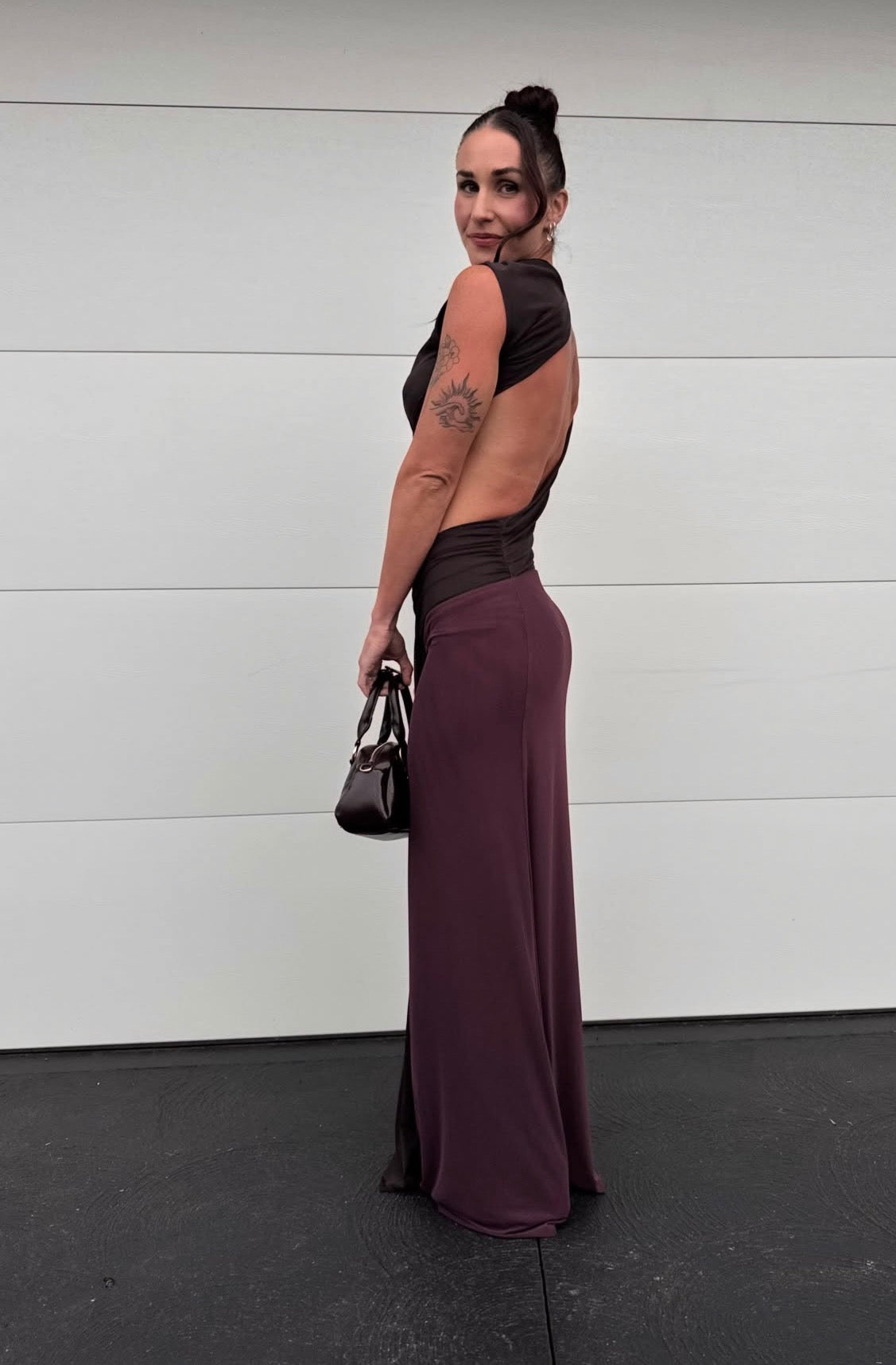 A woman stands in profile wearing a floor-length open-back gown with a black bodice and sleeves and a deep plum-purple skirt, holding a small black handbag against a white paneled wall.