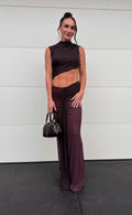 A woman stands against a white paneled wall wearing a dark plum two-piece outfit with a sleeveless high-neck crop top and matching draped wide-leg pants, holding a small dark handbag. - @michalliegovorcin