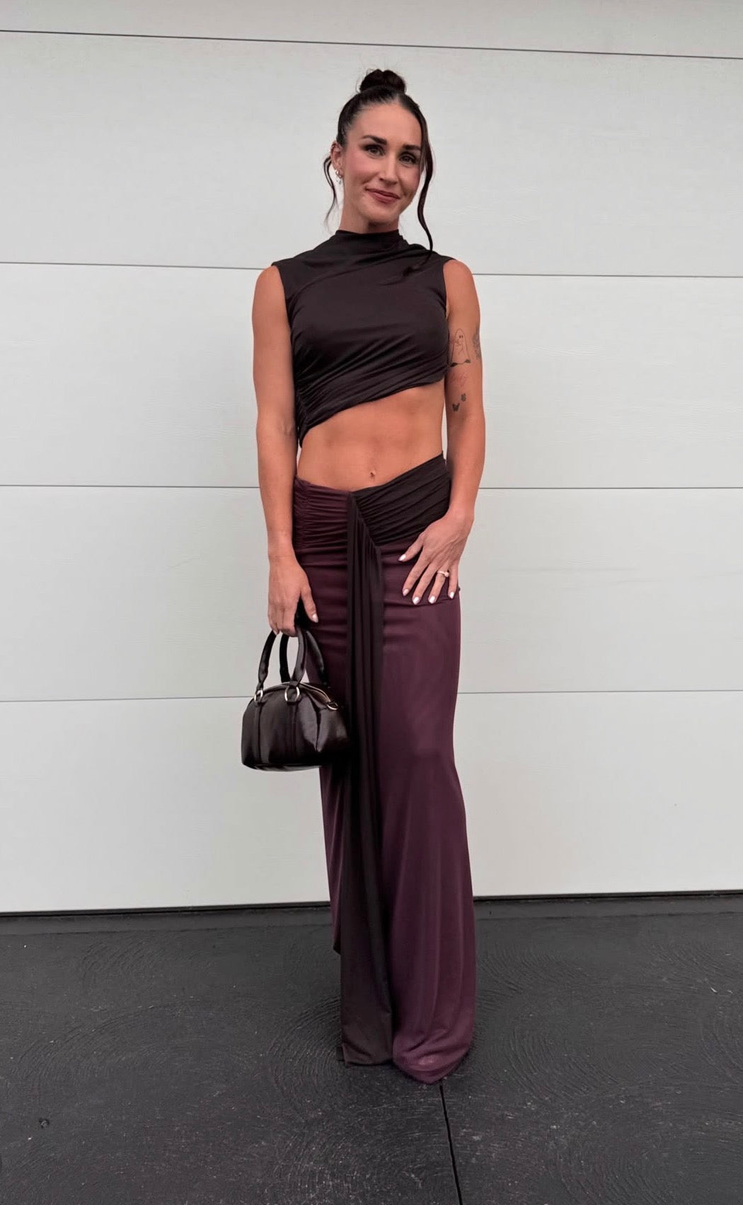 A woman stands against a white paneled wall wearing a dark plum two-piece outfit with a sleeveless high-neck crop top and matching draped wide-leg pants, holding a small dark handbag. - @michalliegovorcin