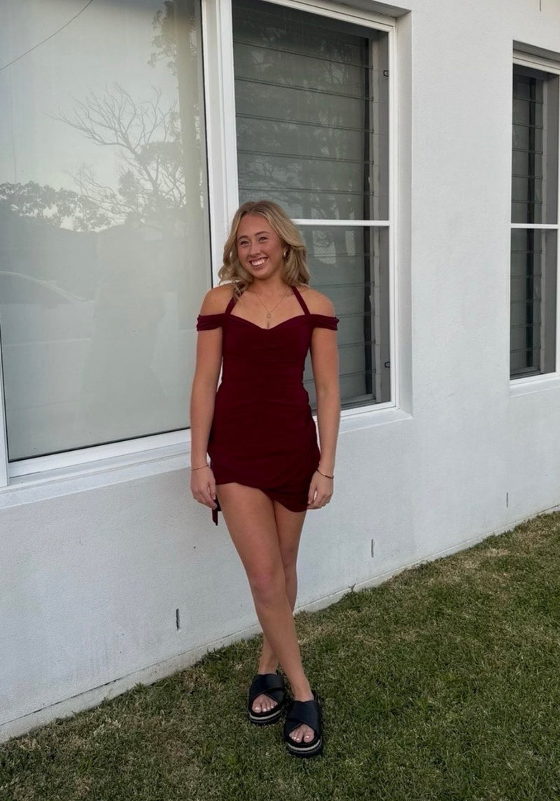 A woman stands outdoors in front of a white house wall and windows, wearing a short off-the-shoulder burgundy mini dress and black slide sandals. - @tahneepotter_