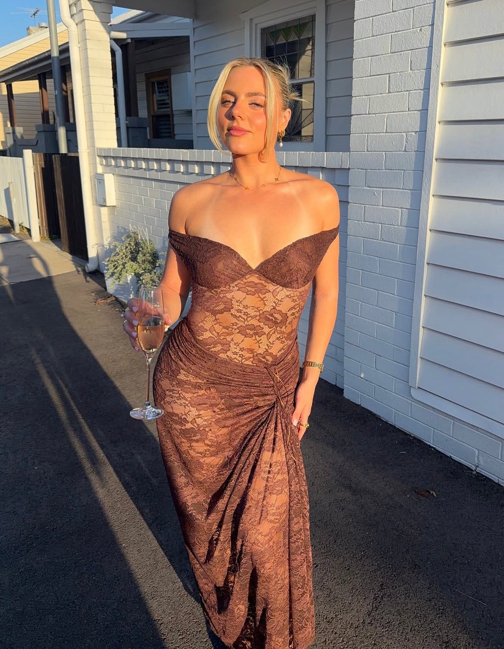 A woman wearing a brown lace off-the-shoulder maxi dress stands outdoors in front of a white house wall, holding a wine glass. - @amityloxley