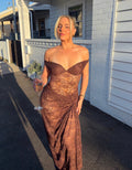 A woman wearing a brown lace off-the-shoulder maxi dress stands outdoors in front of a white house wall, holding a wine glass. - @amityloxley