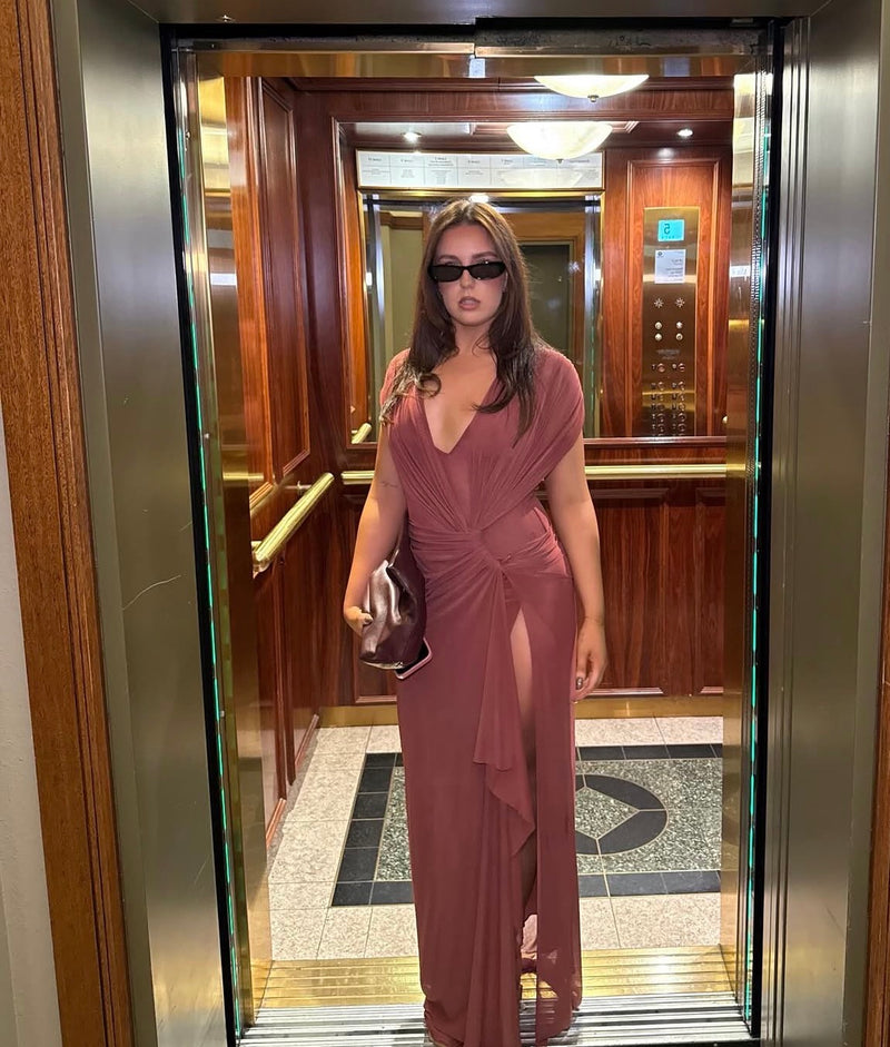 A woman wearing a long, dusty rose wrap-style dress with a thigh-high slit and draped sleeves, standing in an elevator with wood-paneled walls. - @tahliawithers