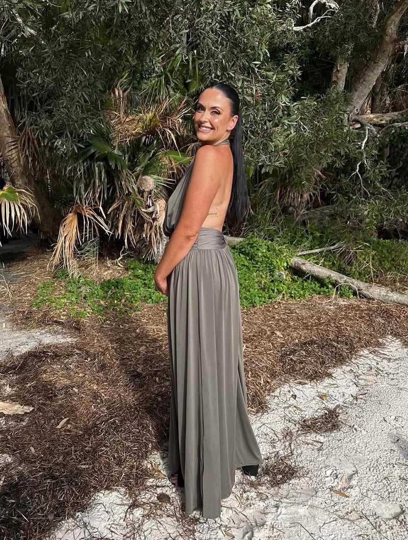 A woman wearing a sleeveless, backless, floor-length taupe-gray maxi dress, standing outdoors on a sandy path with palm trees and green foliage in the background. - @gabypepper