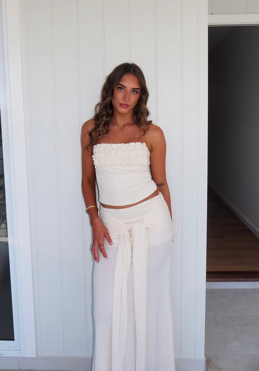 A woman modeling a white two-piece outfit consisting of a strapless cropped top with textured ruffles and matching wide-leg skirt, standing against a white paneled wall. - @violetteclarkk_