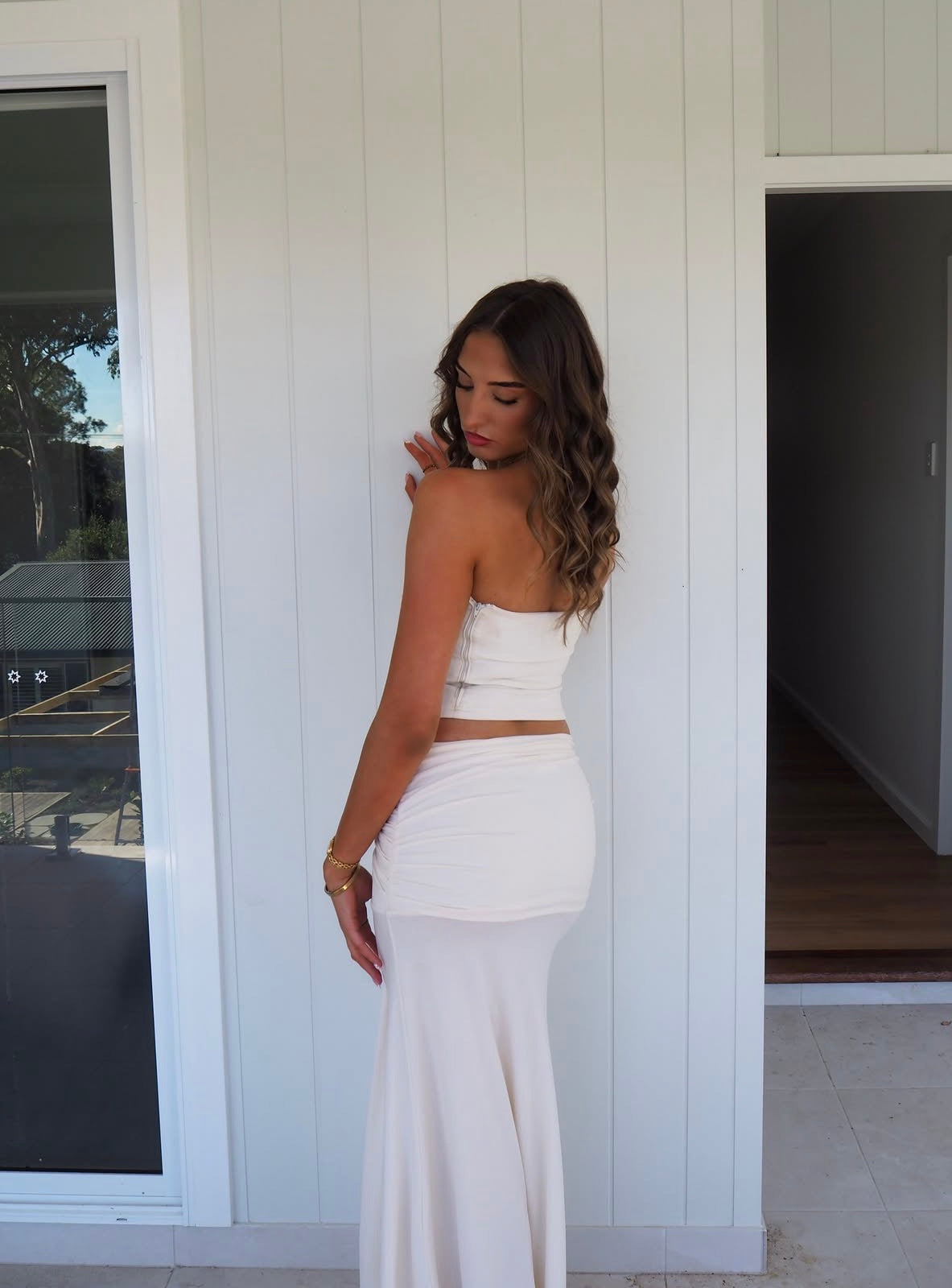 A woman wearing a white strapless tube top and matching white flared skirt, standing in front of a white paneled exterior wall.